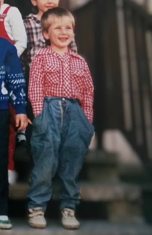 A young boy, Patryk, with a big smile on his face, wearing a red and white checkered shirt tucked into high-waisted blue jeans and sneakers, standing outside with other children partially visible around him.